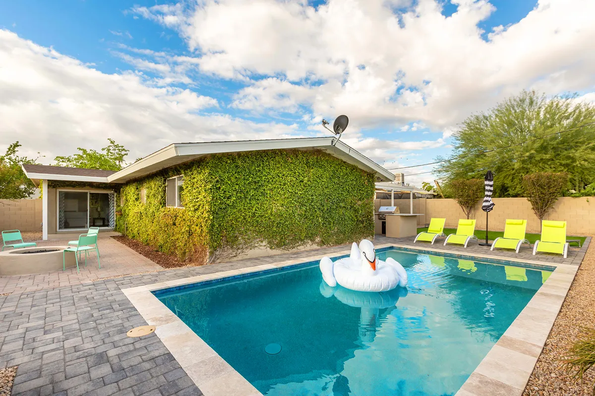 The image features the exterior of a single-family Scottsdale home with a sparkling swimming pool accented by a whimsical swan float. A lush green lawn, inviting lounge chairs, and an ivy-covered wall frame the scene, while the overall mood is playful, inviting, and perfect for outdoor relaxation and gatherings.