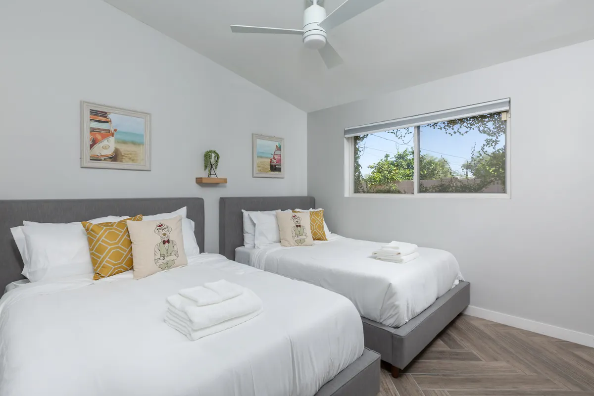 A spacious bedroom featuring two beds with crisp white linens, colorful decorative pillows, modern artwork, and a large window that frames views of lush greenery. The room feels airy and inviting, suited for families or groups.