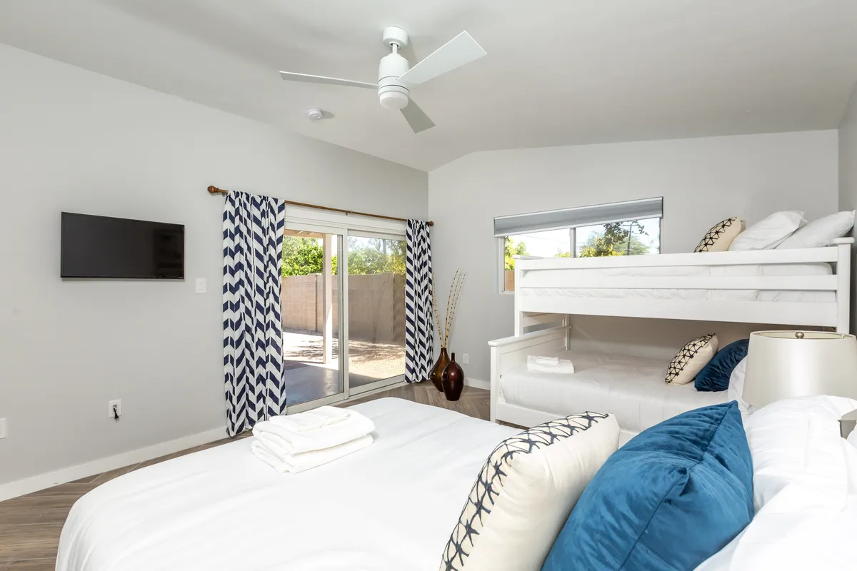 A versatile bedroom setup with a bunk bed and a queen bed, a window with curtains, a television for entertainment, and tasteful decor. The room exudes comfort and is designed for both kids and adults.