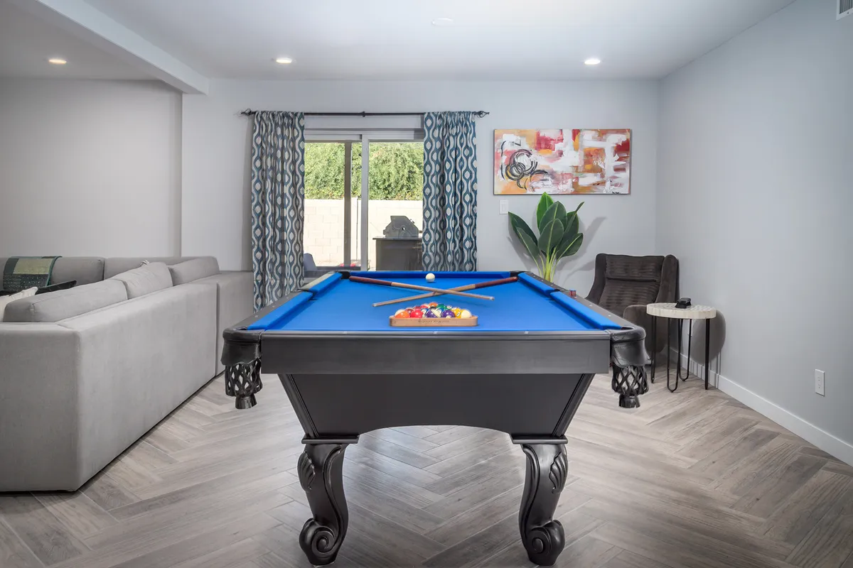 A vibrant, open living area featuring a pool table as a central element, a comfortable sofa, large windows with flowing curtains, colorful artwork on the wall, and a leafy potted plant. The whole scene radiates warmth and activity, with ample space to gather, play, or relax.