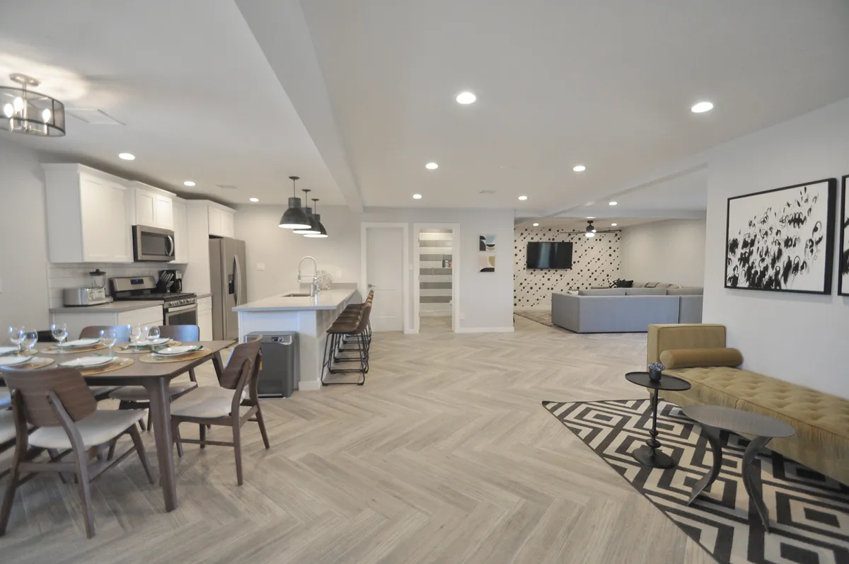 A wide view of an open-plan space featuring a dining table, kitchen island, sofa seating area, and a decorative wall. The scene emphasizes the seamless flow between cooking, dining, and relaxing, all in a bright, modern setting.