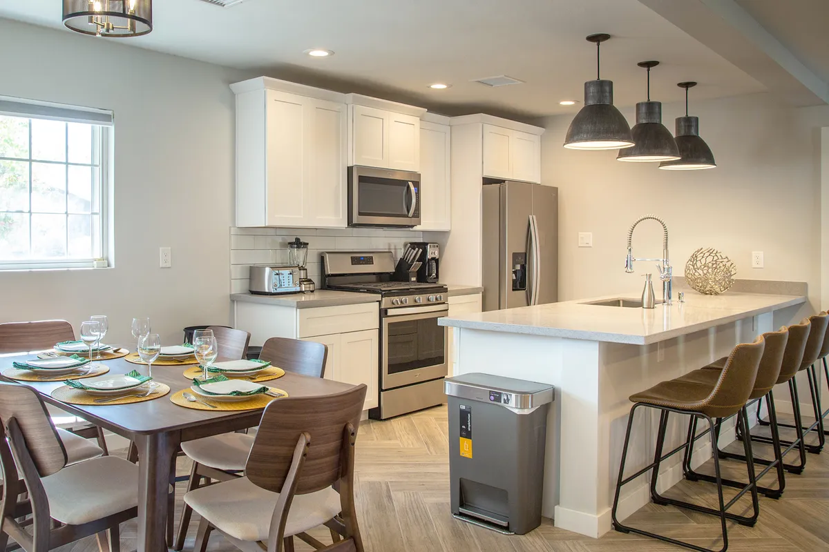 This image showcases a modern, fully equipped kitchen with sleek appliances, a spacious kitchen island with bar stools, and a dining table set for a meal. Pendant lighting fixtures add warmth and style to the space, and the overall atmosphere is inviting and communal, perfect for group gatherings and meal preparation.